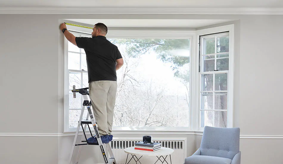 A professional window treatment installer measures a side window in a bay window before installing blinds