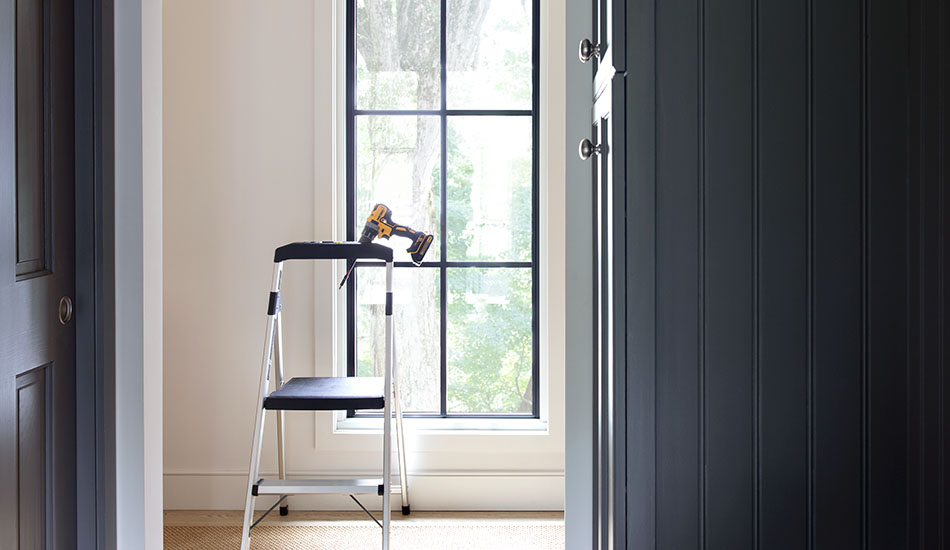 A stepladder with a drill stand in front of a tall narrow window that is bare with no window treatments