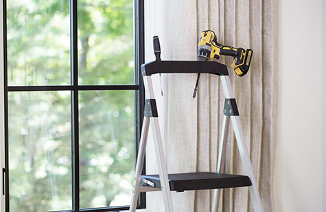 A step ladder with a drill sits in front of a window and shows the first step for how to install shades which is gather tools
