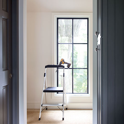 A stepladder sits in front of a tall narrow window in an off-white room with a drill sitting in the holder