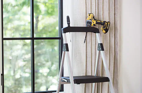 A step ladder sits next to a window with a drill and screwdriver, showing tools needed for how to install drapes