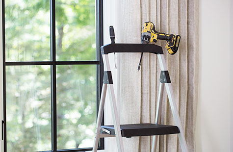 A step ladder and drill in front of a window show some of the tools needed to learn how to install roman blinds