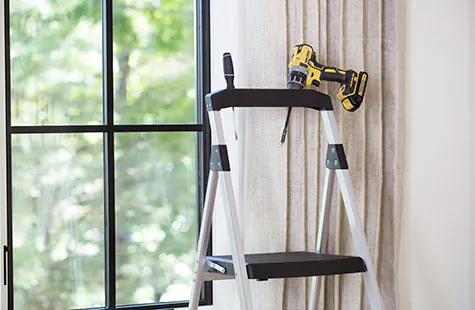 A step ladder and drill in front of a window show some of the tools needed to learn how to install roman shades