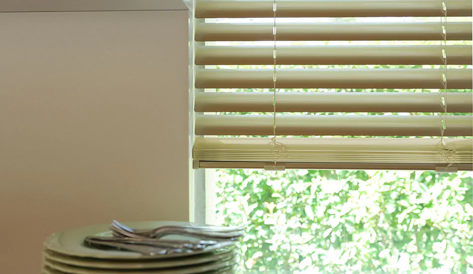 Gold champagne metal blinds used in a kitchen window next to a counter with plates are one option for bay window blinds