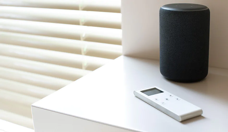 A remote for motorized window treatments sits with a smart home device next to a window with Champagne Metal Blinds