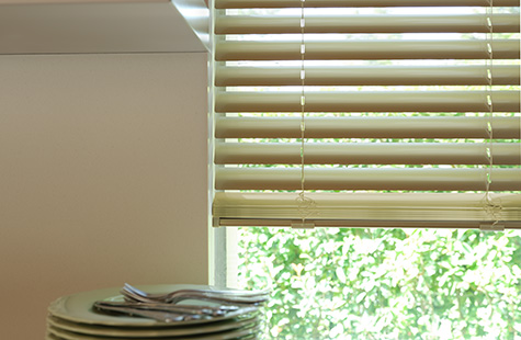 Plates sit on a kitchen countertop in front of a window with Champagne 2-inch Metal Blinds that feature a gold metallic sheen