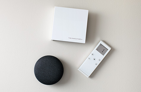 A motorization remote sits on a table next to The Shade Store wireless link and a Google Nest smart home device