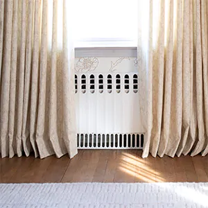 Pinch Pleat Drapery made of Nate Berkus Paloma in Sandstone kisses the warm wood floor of a bedroom with floral wallpaper