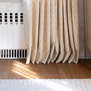 Pinch Pleat Drapery panels made of Paloma in Sandstone gently kiss the warm wood floors which helps hide uneven floors