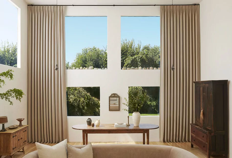 Pinch pleat drapery frame a convivial living room scene with plenty of greenery outside and a couch in the foreground