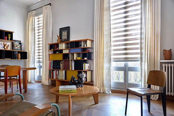 A mid-century modern lounge has Pinch Pleat Drapery made of Raw Silk in Glacier and Double Roller Shades in Windansea Canyon