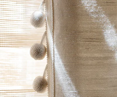 A detail shot of boho curtains of Raw Silk in Glacier feature Samuel and Sons Celine Ombre Ball in Ecru as an embellishment