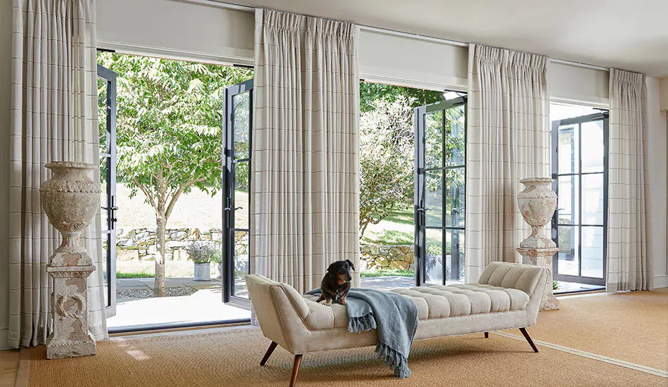 A dog sits on a settee in a large living room with Pinch Pleat Drapery made of Sankaty Stripe in Sand over French doors