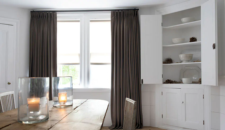 Walnut-colored thermal curtains hanging in a dining room with a vintage wooden table in the foreground with two candles lit