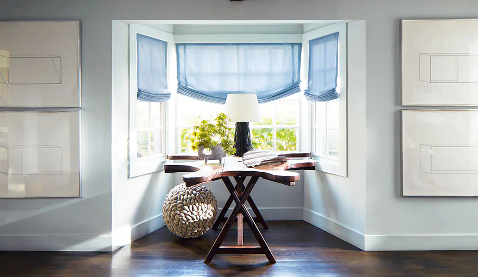 Coastal window treatments of Relaxed Roman Shades made of Andes in Fountain adorn a bay window with a wood table in front