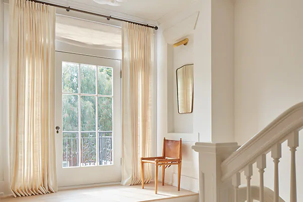 A hallway door has a Relaxed Roman Shade made of Luxe Sheer Linen in Off-White above it and Raw Silk drapery on each side