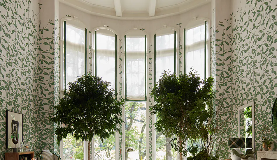 Very tall windows feature flat roman shades made of linen in ivory with green trim in place of bow window curtains