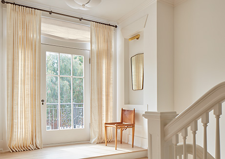 Pinch Pleat Drapery of Raw Silk in Glacier and Relaxed Roman Shades in Luxe Sheer Linen, Off-White adorn a hallway glass door as light-filtering window treatments for privacy and natural light