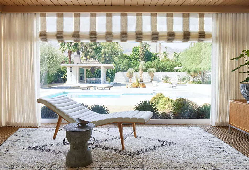 A lounge chair sits in front of a large window with drapes made of Mirador in Ivory and striped Roman Shades