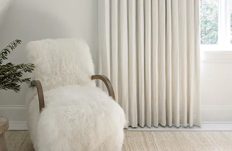 A bright room with a furry chair and light wood floors features off-white curtains that float just above the floor