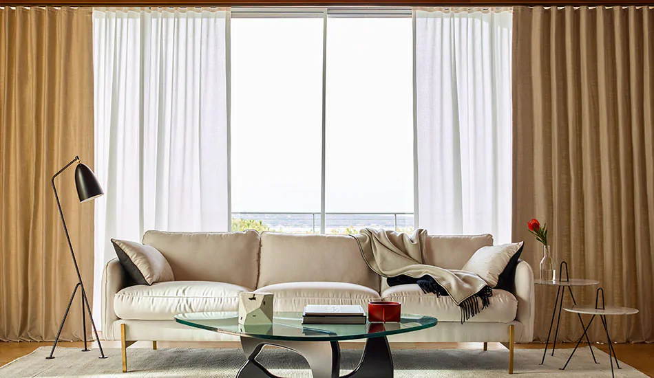 A casual living room features layered curtains made of Ripple Fold Drapery in Neblina, Snow and Alma, Gravel