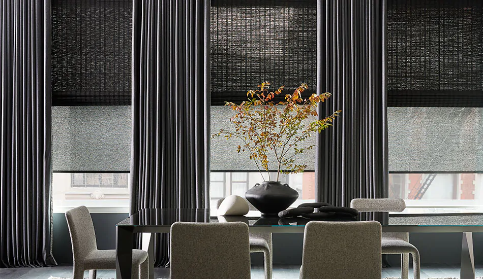 A dining room has Ripple Fold Drapery made of Wool Blend in Charcoal layered with woven and solar shades in shades of gray