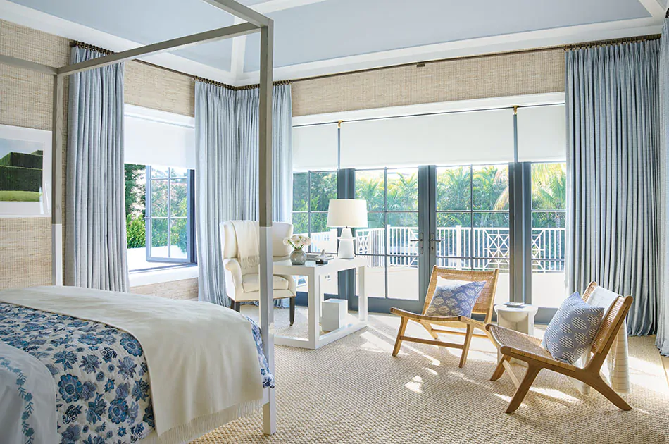 A bedroom has layered window treatments of Tailored Pleat Drapery made of Vanda in Sky and Roller Shades made of Ava in Mist