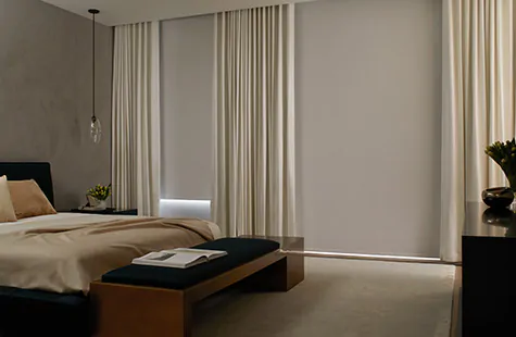 To compare light filtering vs blackout a bedroom has Roller Shades of Cora Blackout in white and blackout lined drapery