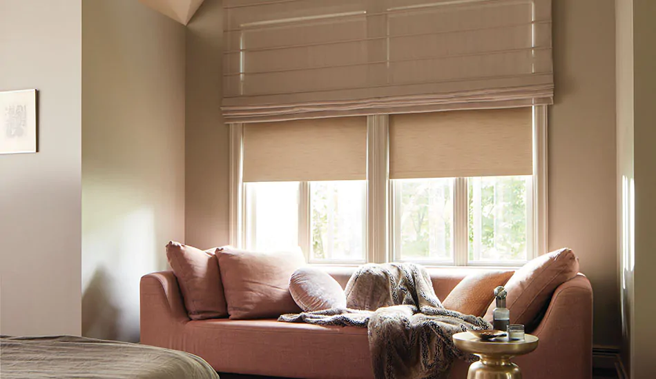 A warm, inviting sitting area features a salmon couch with pillows and layered Aventura Roman Shades and Roller Shades