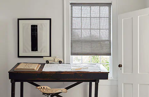 Bamboo blinds made of Roller Shades in Jackson, Sand, cover a small window in an office with a dark wood table