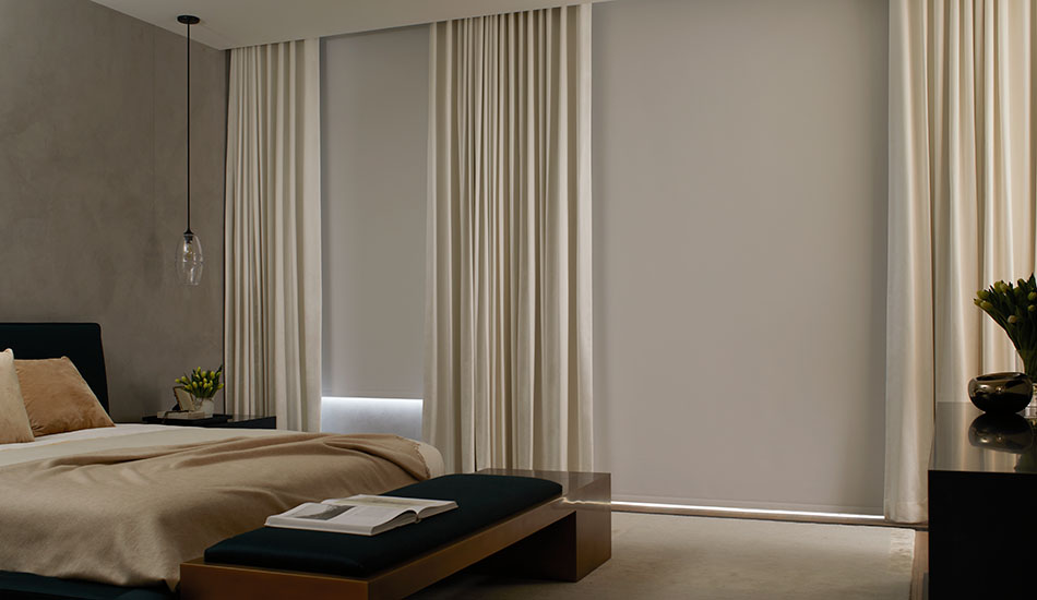 A modern bedroom is darkened thanks to Roller Shades made of Cora Blackout and Ripple Fold Drapery with blackout lining