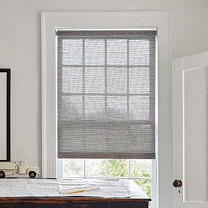 A small window in a rustic office with a wood desk features a Roller Woven Wood Shade made of Jackson in Sand