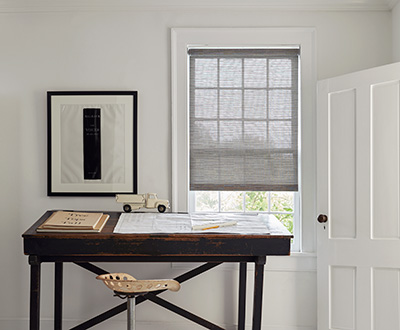 Woven shades made of Jackson in Sand offer a light grey color to a small office with a dark wood desk