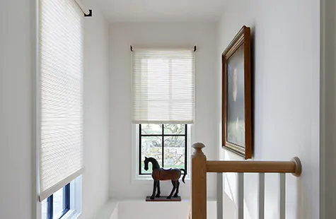 A stairwell with a wood horse decor piece has high windows with woven Roller Shades made from Mesa Verde material in Sand