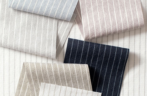 Windsor Stripe fabric in different colors is folded on a table and offer a subtle, sophisticated look for linen Roman Shades
