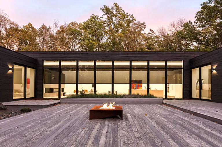 A modern home has solar shades at night that you can see through from the large deck with a firepit that abuts tall windows