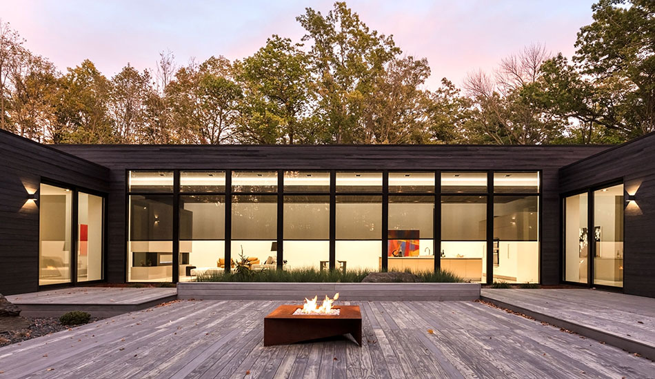 An exterior of a modern home with a wooden patio and a fire pit with a long expanse of solar shades for windows