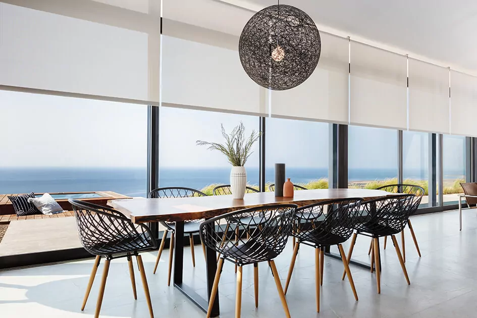 A coastal home features solar shades for sliding glass doors over the floor-to-ceiling glass panels with a view of the sea