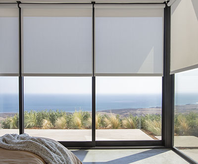 Multiple shades pulled to the same height in the corner of a sunny modern bedroom with coastal views of the beach below