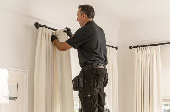 A professional window treatment installation hangs Tailored Pleat Drapery in a warm, neutral-colored living room