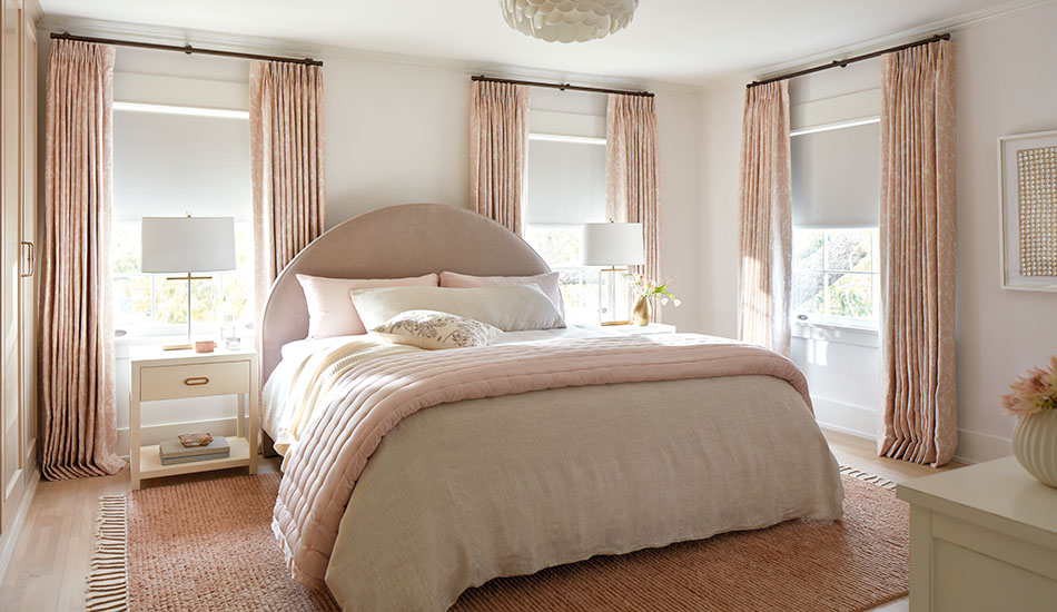 An inviting bedroom features Tailored Pleat Drapery made of Chinoiserie in Blush and Roller Shades made of Ava in Birchwood