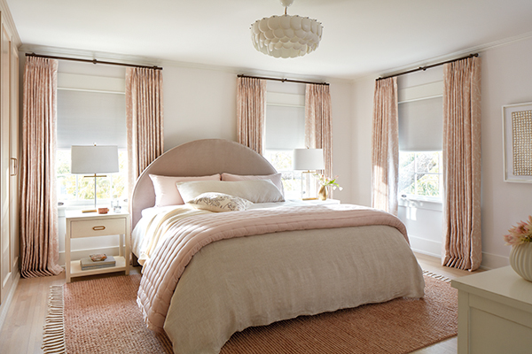 A cozy bedroom with pink and white colors has blackout Roller Shades for windows made of Ava in Birchwood paired with drapery