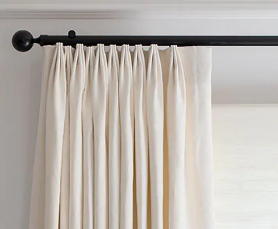 A close-up shot of oyster-colored thermal curtains, specifically tailored pleat drapery, hanging along a black rod