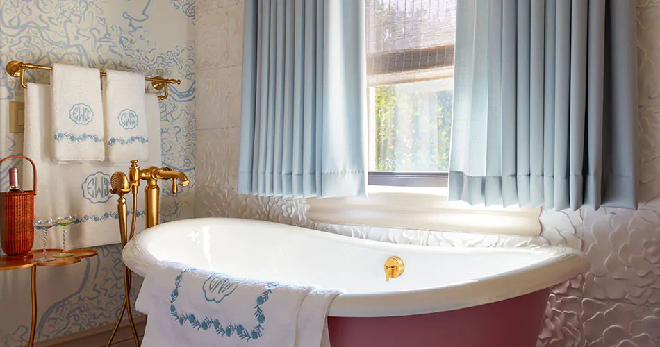 A luxe bathroom with blue and white patterned wallpaper and a pink tub has Tailored Pleat Drapery made of Andes in Fountain
