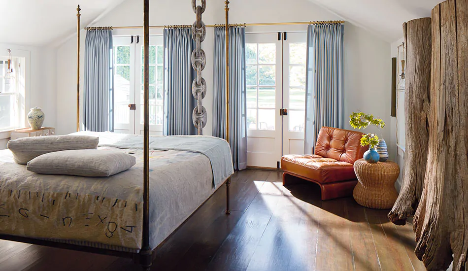 A bedroom has rustic wood and metal tones with coastal window treatments of Tailored Pleat Drapery made of Andes in Lake