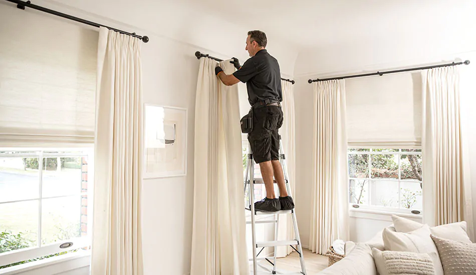 A professional installer stands on a step ladder in a living room showing how to install curtain rods and hang the drapery