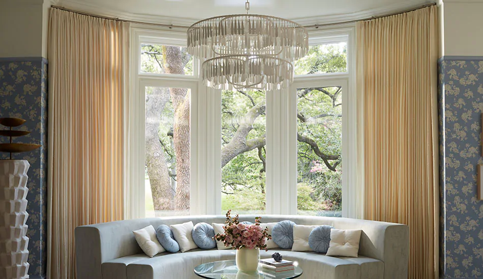 Tailored pleat drapery is used as bow window curtains that hang on a curved rod in a luxurious room with a chandelier