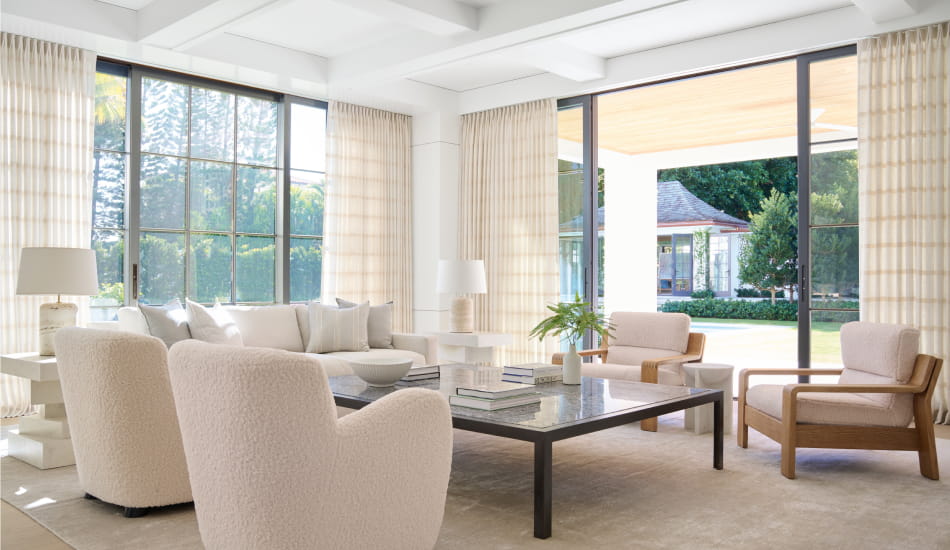 Corner of a bright, airy living room with windows featuring Tailored Pleat Drapery in Victoria Hagan's Lily fabric in Buff.