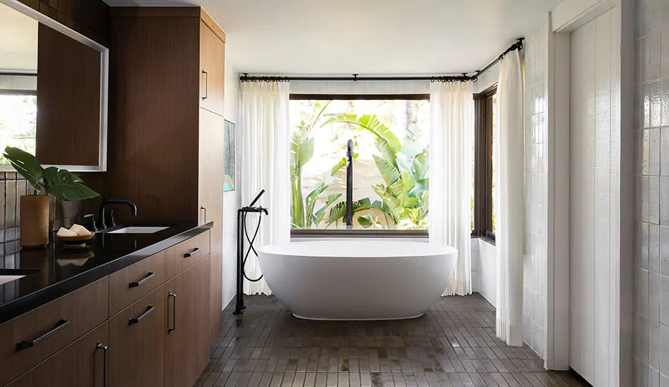 A rustic bathroom with a large white tub and warm wood cabinets features corner window curtains made of Linen Sheers in White