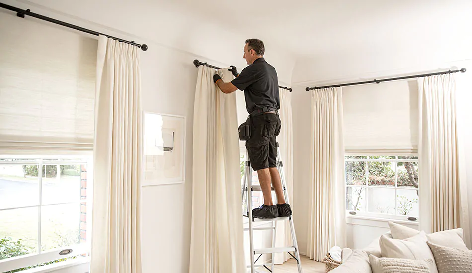 A professional installs off-white toned curtains high and wide on black rods showing how to hang curtains